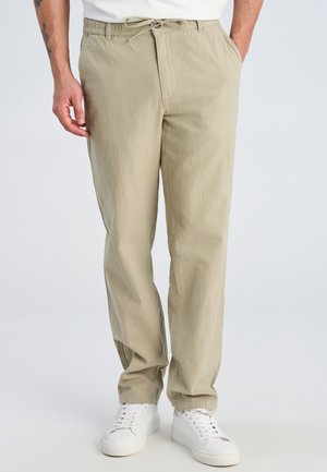 Mężczyzna w beżowych spodniach ze ściągaczem i białych trampkach stoi na tle jednolitego tła, z jedną ręką w kieszeni.