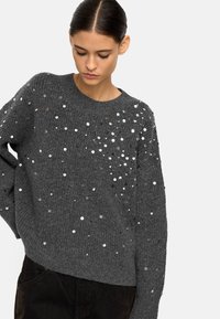 Szary sweter dzianinowy z okrągłym dekoltem, zdobiony rozrzuconymi srebrnymi cekinami dla uzyskania faktury i detali. Luźny krój z długimi rękawami.