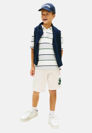 Ragazzo giovane che indossa una polo a righe, pantaloncini bianchi, scarpe da ginnastica bianche, cappellino blu navy e maglione blu navy drappeggiato sulle spalle, sorridente e in piedi.