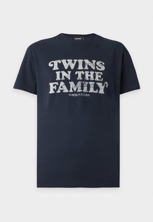 Junger Mann steht mit einer Hand in der Tasche, trägt ein schwarzes "Twins In The Family" T-Shirt, abgenutzte blaue Jeans und schwarze Sneaker vor weißem Hintergrund.