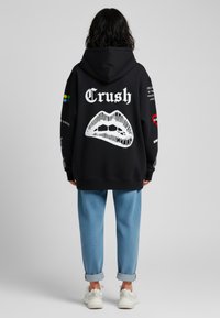 Felpa nera con grafica di labbra bianche e testo "Crush" sulla schiena. Abbinata a pantaloni blu chiaro con risvolti e scarpe da ginnastica bianche.