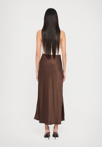 Brown satin maxi skirt with side slit, smooth texture, high waist, and a flowing design. Worn with a simple white top and heeled sandals.