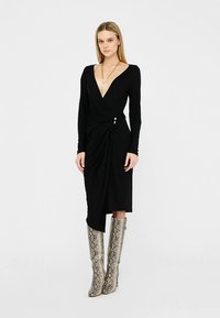 Schwarzes Wickelkleid mit langen Ärmeln, tiefem V-Ausschnitt, drapierten Details an der Taille und einer dekorativen Akzentuierung. Kombiniert mit grauen kniehohen Stiefeln mit Schlangenprint.