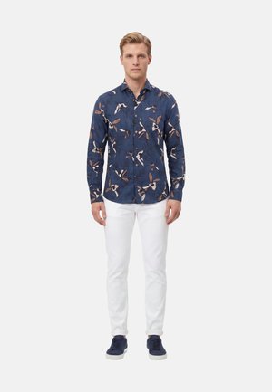 Uomo che indossa una camicia blu a fiori a maniche lunghe con bottoni, pantaloni bianchi e sneakers blu scuro, in piedi su uno sfondo semplice.