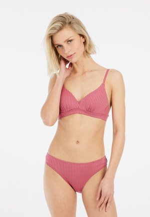 Vrouw met kort blond haar, gekleed in een geribde oudroze beha en bijpassende onderbroek, poserend tegen een witte achtergrond.