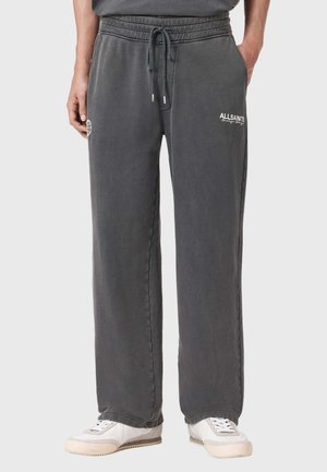 Mężczyzna w luźnych, ciemnoszarych spodniach dresowych z troczkiem w pasie i białych sportowych butach, stojący na jednolitym jasnym tle.