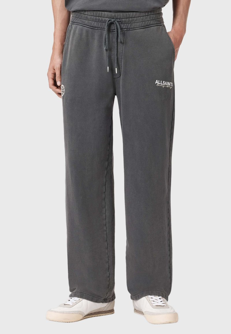 Άνδρας που φοράει φαρδιά σκούρα γκρι φόρμα με κορδόνι στη μέση και λευκά αθλητικά παπούτσια, στέκεται μπροστά σε απλό ανοιχτό φόντο.