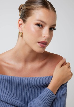 Gouden oorring in hartvorm gedragen door een model. Het model draagt een blauwe geribbelde off-shoulder trui. De huid heeft een gladde textuur en de nagels zijn neutraal.