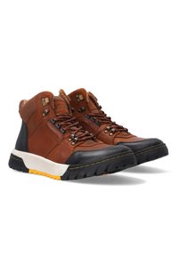 Bruine leren wandelschoenen met een zwarte rubberen neuskap, gele zoolaccenten, metalen oogjes en netdetails aan de zijkanten.