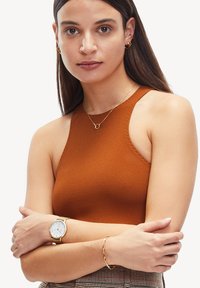 Rustfarbene ärmellose Bluse aus strukturiertem Stoff, kombiniert mit einer goldenen Uhr und einem Armband, die ein schlichtes und elegantes Design aufweisen.