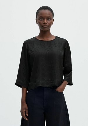 Une femme présente un chemisier noir texturé à manches trois-quarts et un pantalon foncé sur un fond clair uni.