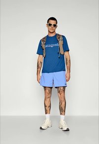 Mėlyna marškinėliai su "Patagonia" logotipu, šviesiai mėlynos šortai, smėlio spalvos sportbačiai ir chaki kuprinė. Modelis turi tatuiruotes ir saulės akinius.