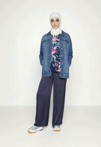 Jaqueta de ganga sobre uma blusa de estampado floral, calças largas azul-marinho e ténis azul claro com sola laranja. O hijab branco completa o visual.