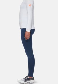 Camicia bianca a maniche lunghe con logo arancione, abbinata a leggings blu scuro con motivo geometrico e superficie testurizzata. Scarpe da ginnastica bianche e grigie.