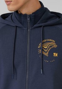 Granatowa rozpinana bluza z kapturem z wysokim kołnierzem, ozdobiona złotym logo i sznurkami. Gładka tkanina z prążkowanymi wykończeniami wokół szyi.