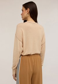 Maglione in maglia beige con spalle scese, maniche lunghe e una vestibilità rilassata, abbinato a pantaloni marroni con strisce laterali. Texture morbida.