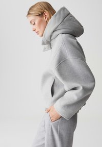 Frau im Profil, die einen hellgrauen, oversized Pullover mit großem gepolstertem Kragen und passenden grauen Hosen trägt, die Hand in der Tasche, vor einem weißen Hintergrund.