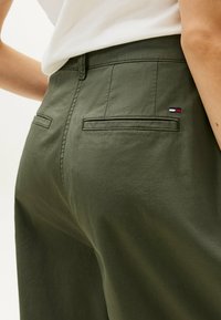 Olijfgroene broek met een gladde textuur, voorzien van twee achterzakken en een klein geborduurd logo in rood, wit en blauw op de tailleband.
