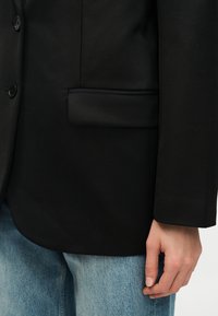 Μαύρο σακάκι με μπροστινή τσέπη με καπάκι και κουμπιά, φορεμένο πάνω από ανοιχτό μπλε τζιν, με ένα χαλαρό χέρι στο πλάι.