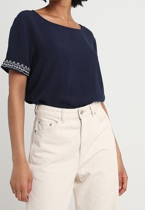 Haut bleu marine à manches courtes avec un col carré et un détail brodé blanc sur les manches, associé à un jean clair taille haute.