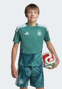 Equipamento de futebol verde da Adidas, composto por uma camisola de manga curta com padrão gráfico e calções combinando, segurando uma bola de futebol multicolorida.