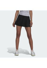 Falda de tenis negra en línea A con un diseño en capas, hecha de material transpirable. Combinada con zapatillas deportivas blancas con acentos rojos.