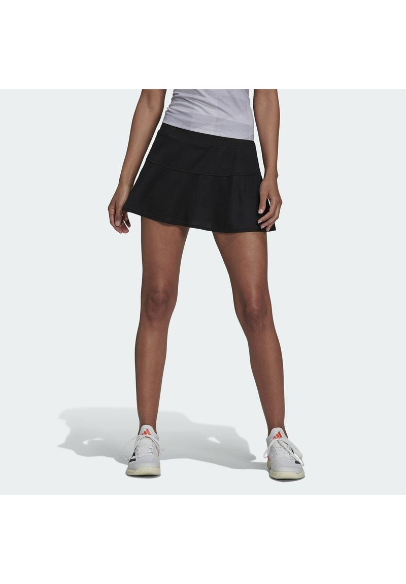 Falda de tenis negra en línea A con un diseño en capas, hecha de material transpirable. Combinada con zapatillas deportivas blancas con acentos rojos.