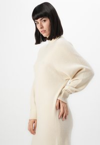 Donna con capelli scuri e lisci, vestita con un abito lungo in maglia color crema a maniche lunghe, in piedi con una mano sui fianchi su uno sfondo bianco.