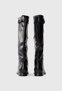 Um par de botas altas de couro pretas com zíperes laterais e tiras no tornozelo, vistas por trás em um fundo branco liso.