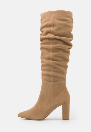 Bottes hauteur genou à bout pointu en daim beige, dotées d'un design froncé et d'un talon bloc. Texture lisse avec une finition sans couture.