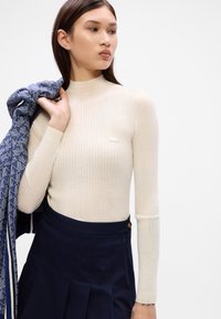 Sweater creme canelado com gola alta e mangas longas, emparelhado com uma saia plissada azul-marinho com um detalhe de botão.