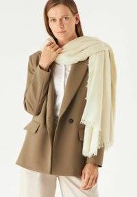 Beige wollen sjaal met franjes hangt over een lichtkleurig shirt, gedragen door een persoon in een bruin colbert tegen een neutrale achtergrond.