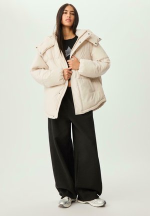 Giacca puffer beige con collo alto e chiusura con zip, abbinata a pantaloni ampi neri e sneaker argentate. Texture liscia e trapuntata.