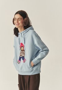 Világoskék kapucnis pulóver elején egy grafikával, amin egy csíkos pulóvert, rózsaszín pomponos sapkát és kék nadrágot viselő medve látható; húzózsinórral és első zsebekkel ellátva.