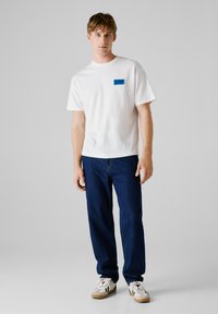 Witte katoenen t-shirt met een blauw vierkant logo op de linkerkant van de borst, gecombineerd met donkerblauwe denim jeans en beige sneakers met zwarte accenten.
