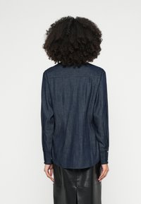 Tmavá denimová košeľa s dlhými rukávmi, zapínanými manžetami, detailom na zadnom dielci a rovného strihu. Kombinovaná s elegantnou čiernou koženou sukňou.