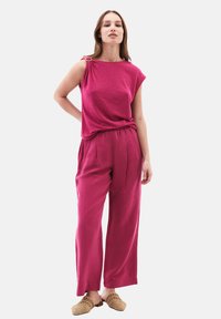 Vaaleanpunainen lyhythihainen paita, jossa on olkapään korostus, yhdistettynä samanvärisiin leveälahkeisiin housuihin ja käytettynä vaaleanruskeilla liukujalkineilla. Silkkinen pinta.