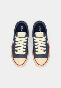 Zapatillas Converse All Star de color azul marino con cordones blancos, punteras beige, ribetes rojos y suelas blancas, vistas desde arriba sobre un fondo blanco.