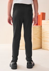 Mustat, istuvaksi leikatut housut, joissa on sileä pinta, joustava vyötärönauha ja yksi takatasku, yhdistettynä kiiltäviin mustiin kenkiin.