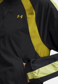 Zwarte windbreakerjas met een gele accent op de schouders en mouwen, voorzien van een ritszak en elastische manchetten. Onder Armour-logo zichtbaar.