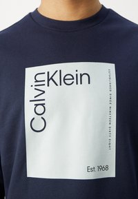 Personne portant une chemise bleu marine avec un grand carré blanc et le texte "Calvin Klein", ainsi que le détail "Établi depuis mille neuf cent soixante-huit".