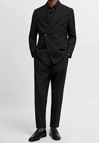 Czarny dwurzędowy garnitur o dopasowanym kroju, z wyciętymi klapami, bocznymi kieszeniami oraz pasującymi czarnymi mokasynami. Gładka tkanina i strukturalny design.