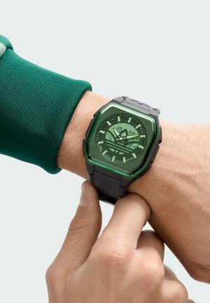 Persona ajustando un reloj digital-analógico negro con cara cuadrada de tono verde en la muñeca, vistiendo una camiseta de manga larga verde.