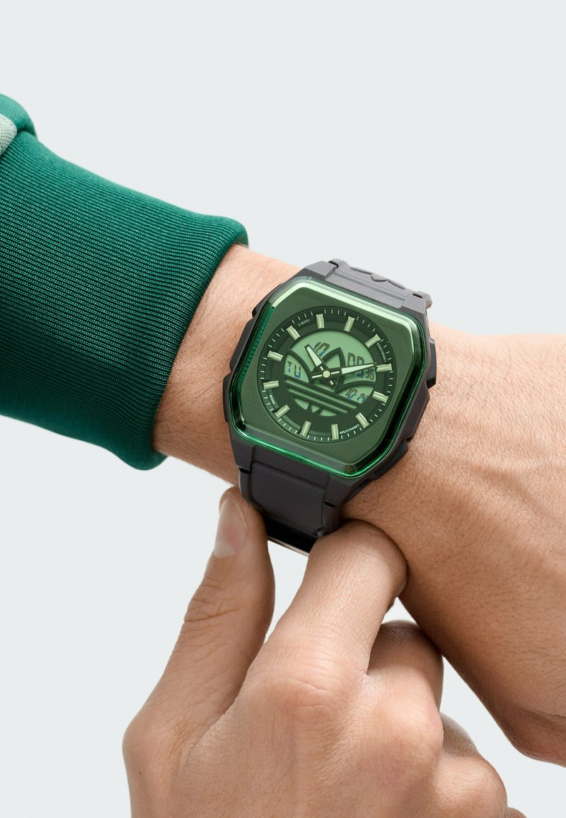 Persoon die een zwart digitaal-analoog horloge met een groen getinte vierkante wijzerplaat op de pols verstelt, gekleed in een groen shirt met lange mouwen.
