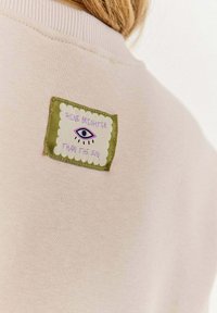 Světle růžová mikina s vyšitým zeleným obdélníkovým nášivkem, na kterém je grafika oka a text „SVÍTĚJ JASNĚJI NEŽ SLUNCE.“