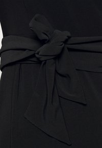 Černé látkové šaty s uzlovým páskem v oblasti pasu, hladkou texturou a vypasovaným střihem.