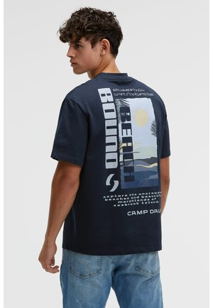 Junger Mann mit lockigem Haar, trägt ein marineblaues T-Shirt mit der Aufschrift "Beach Bound" und hellblaue Jeans, steht seitlich und blickt über die Schulter zurück.