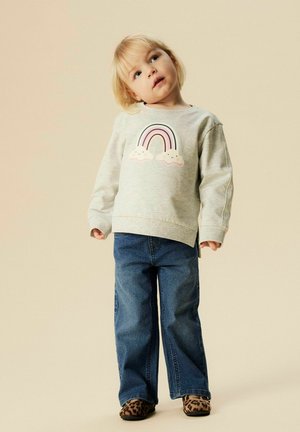 Toddler wearing a light gray sweatshirt with a rainbow and clouds graphic, blue jeans, and leopard print shoes, standing on a beige background.