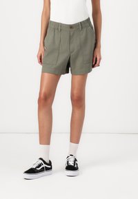 Khakishorts med knappstängning och elastisk midja, med två framfickor. Kombinerat med svarta sneakers och vita strumpor.