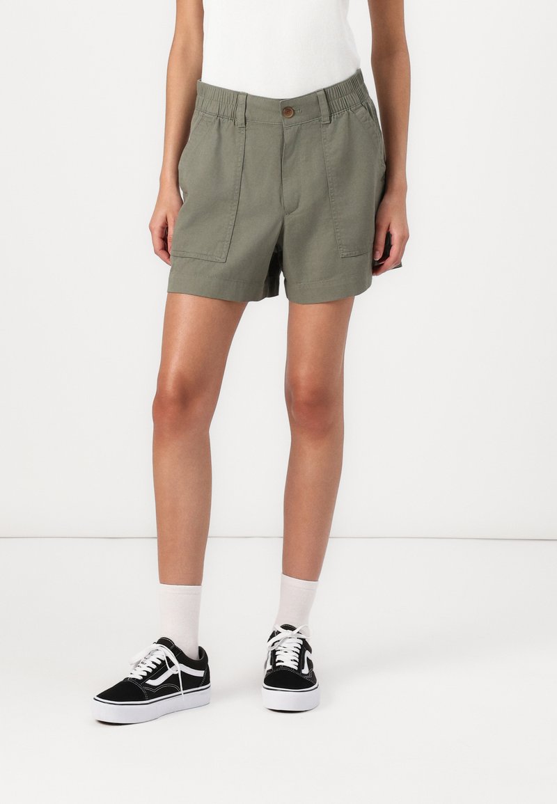 Khakishorts med knappstängning och elastisk midja, med två framfickor. Kombinerat med svarta sneakers och vita strumpor.